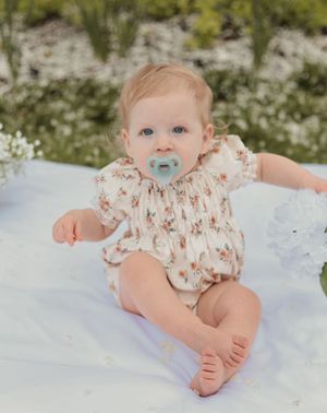 Cute baby girl in floral romper with pacifier sitting on white blanket among wildflowers.