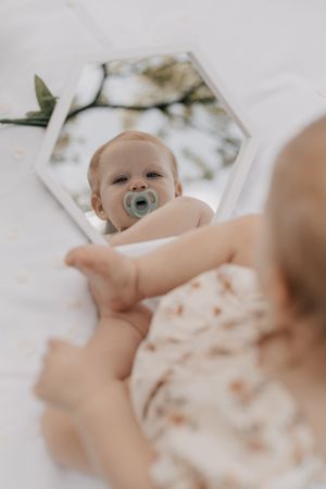 A cute baby with a pacifier gazes at their reflection in a hexagonal mirror outdoors.
