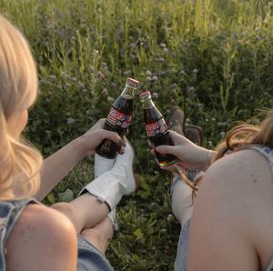 Two women clinking Dr Pepper bottles while sitting together in a wildflower meadow at sunset.