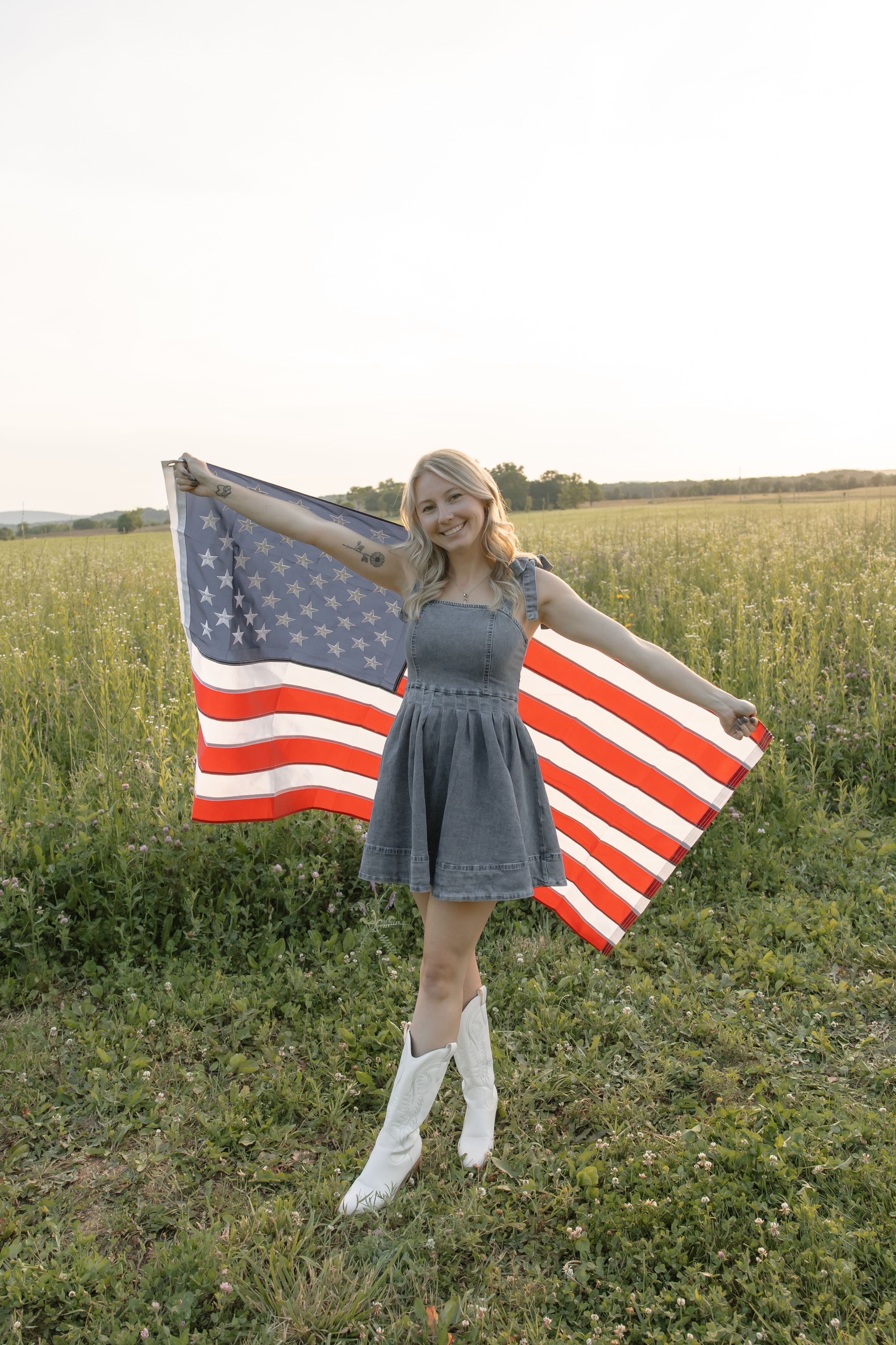 Smiling blonde woman in denim dress holding American flag wide open in green field at sunset.