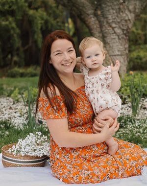 Smiling mother in orange floral dress holds her adorable baby girl in a garden with white wildflowers.