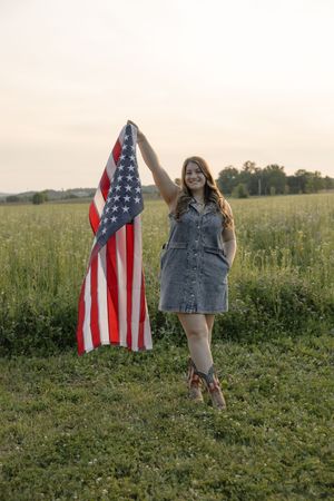 Smiling woman in denim dress holding American flag in green field at golden hour sunset.