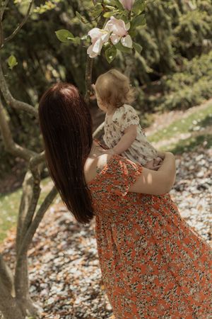 Mother in floral orange dress holding baby near blooming magnolia tree in garden.