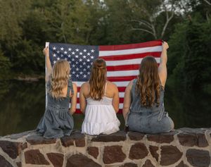 Three women sit on a stone wall holding an American flag with a serene river and trees behind them.