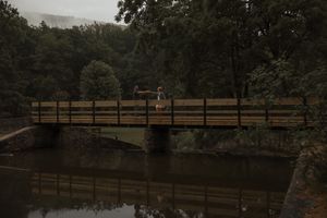 Couple standing on a rustic wooden bridge surrounded by misty green forest reflected in calm water.