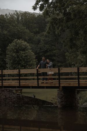 Couple embracing on a wooden bridge over a calm river surrounded by lush green trees on overcast day.