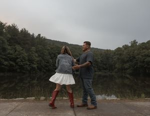 A couple holds hands by a serene lake surrounded by lush green forest.