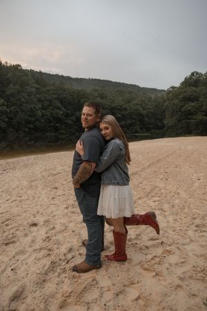 Couple embracing on sandy riverbank at dusk, woman in white dress and red boots.