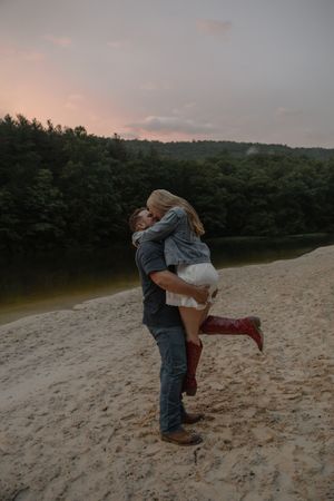 Man lifts and embraces woman in red boots on a sandy riverbank surrounded by lush green forest at dusk.