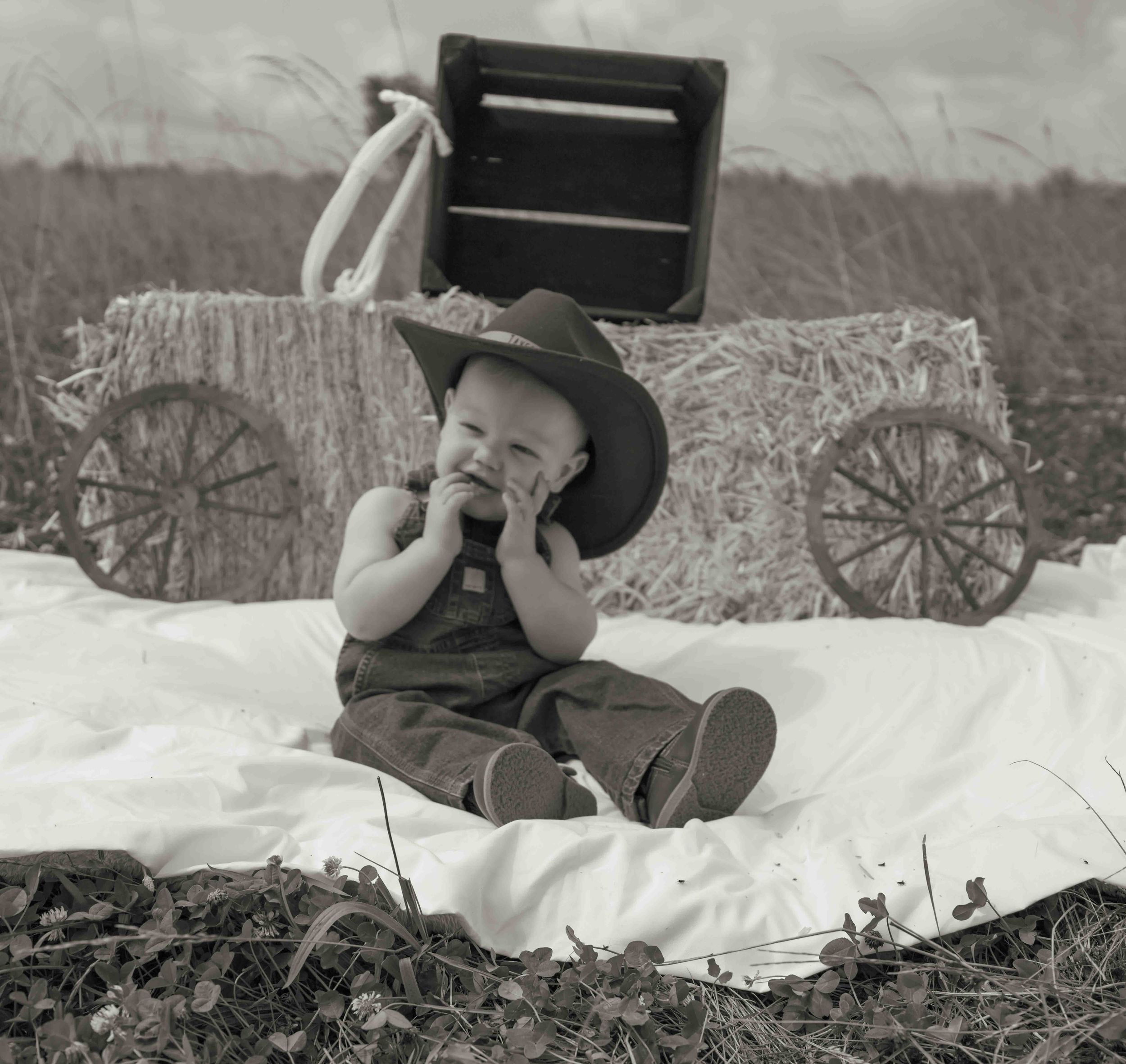 Black and white photo of baby in cowboy hat sitting on blanket near hay bales in field.