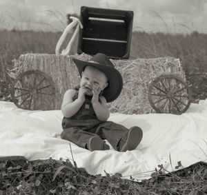 Black and white photo of baby in cowboy hat sitting on blanket near hay bales in field.
