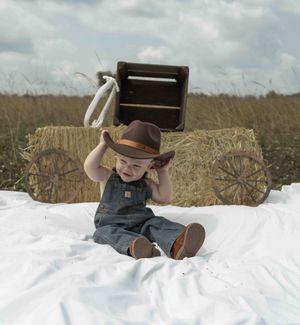 Adorable baby in cowboy hat and overalls sitting on white blanket in rustic farm field.