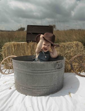 Laughing baby wearing cowboy hat sitting in galvanized tub with hay bales and wagon backdrop.