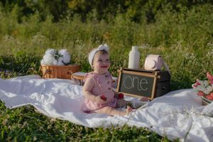 Baby girl in pink dress celebrating first birthday on white blanket in sunny outdoor garden setting.