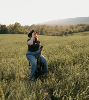 A woman in a black cowboy hat sits with a guitar in a sunlit green field.