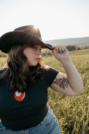 A tattooed woman in a black tee and cowboy hat poses in a sunlit green field.