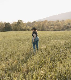Woman in cowboy hat and jeans stands alone in a vast green field with autumn trees and mountains behind her.