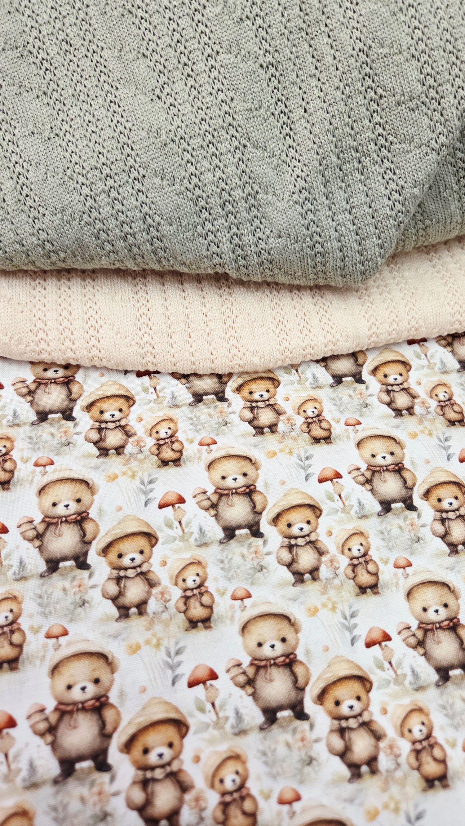 Stacked baby fabrics featuring cute teddy bear mushroom print,soft cream knit,and sage green textured blanket layers.