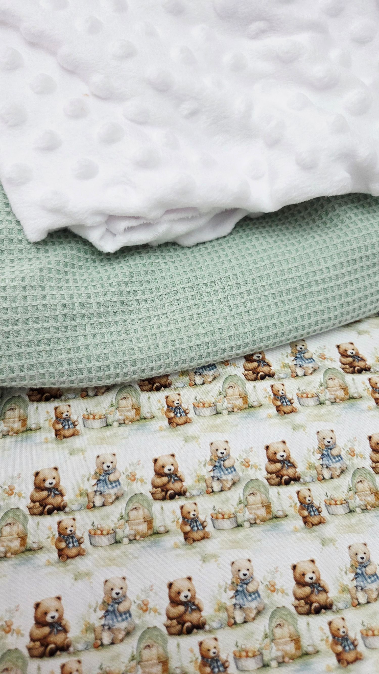 Stacked fabrics featuring teddy bear print, sage green waffle texture, and white minky dot material for sewing projects.