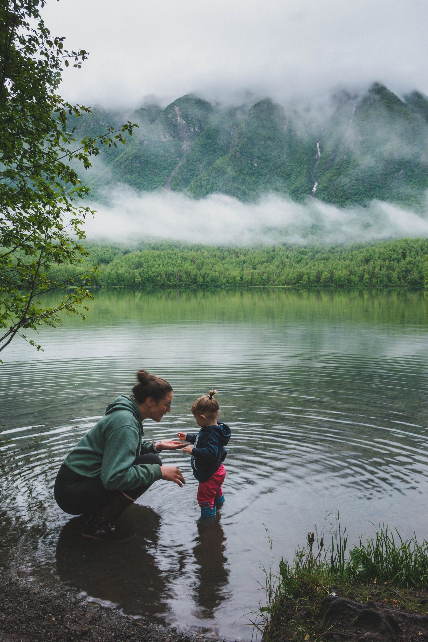 Mother and toddler playing at misty mountain lake shore in lush green landscape.
