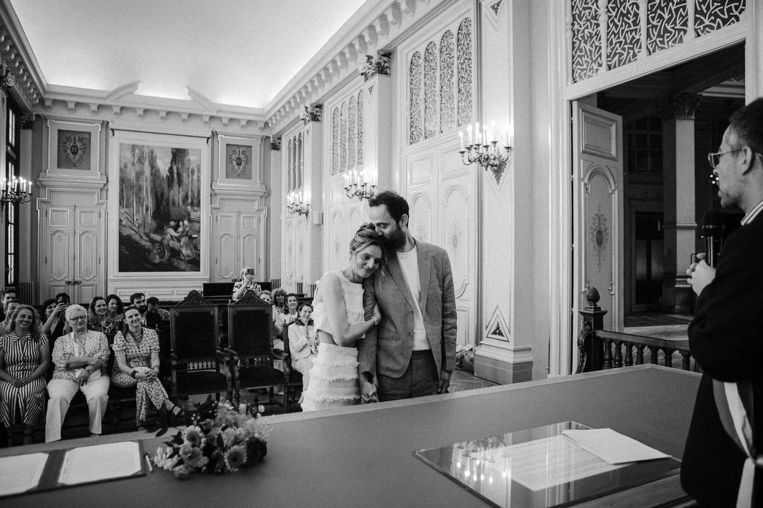 Black and white wedding ceremony photo of couple embracing at altar in elegant ornate hall with seated guests.