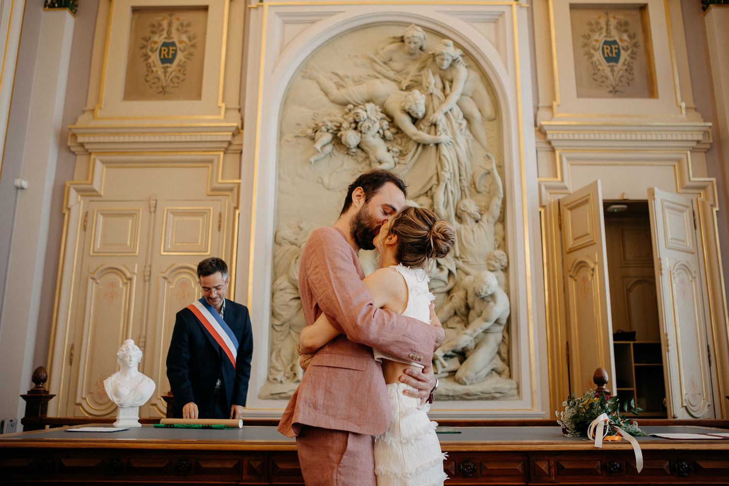 Couple shares first kiss at French civil wedding ceremony with ornate baroque sculpture and official in tricolor sash.