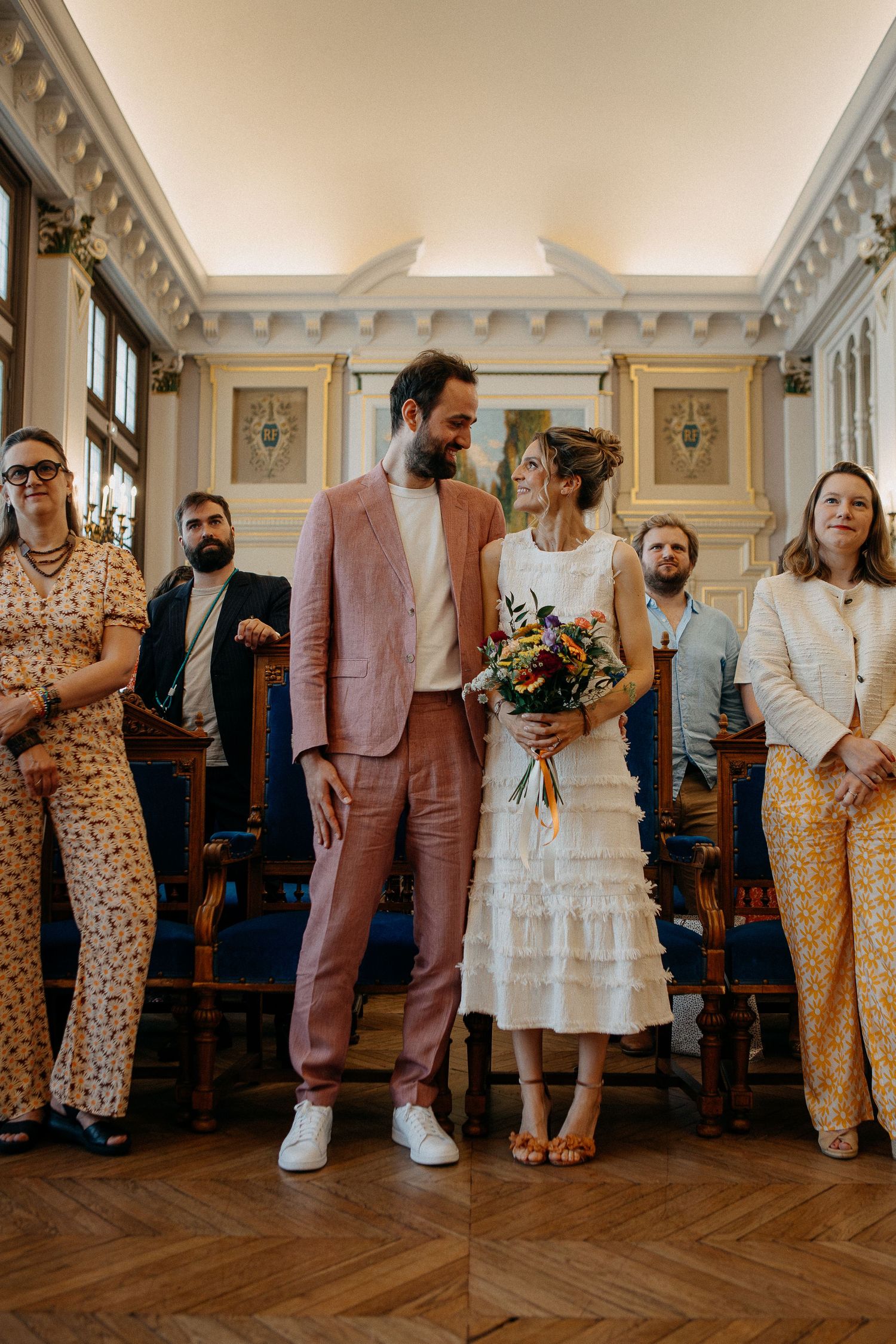 Bride and groom smiling together at civil wedding ceremony, bride holding colorful bouquet, guests standing beside them.