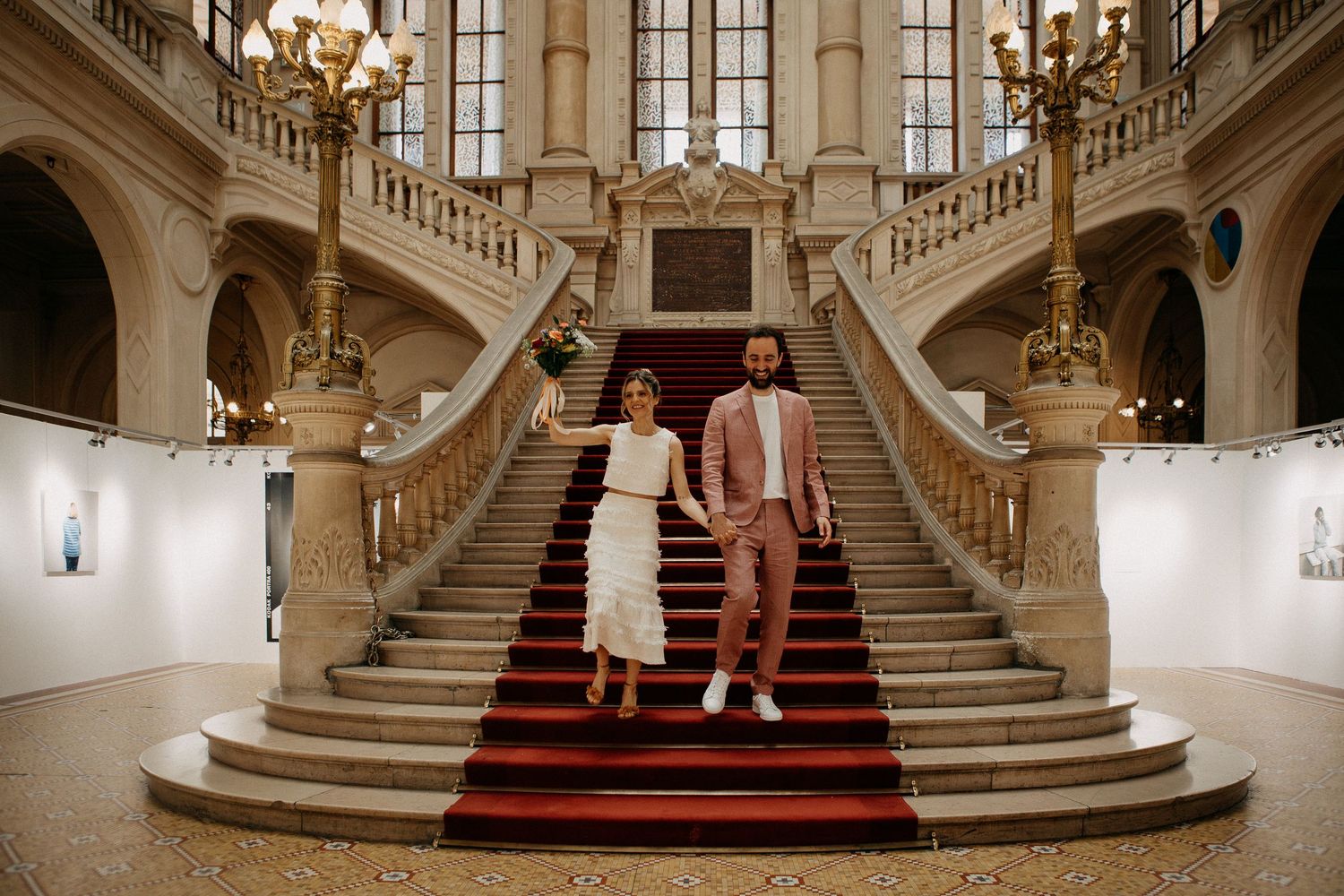 Couple in wedding attire holding hands on red-carpeted grand staircase in elegant historic building.