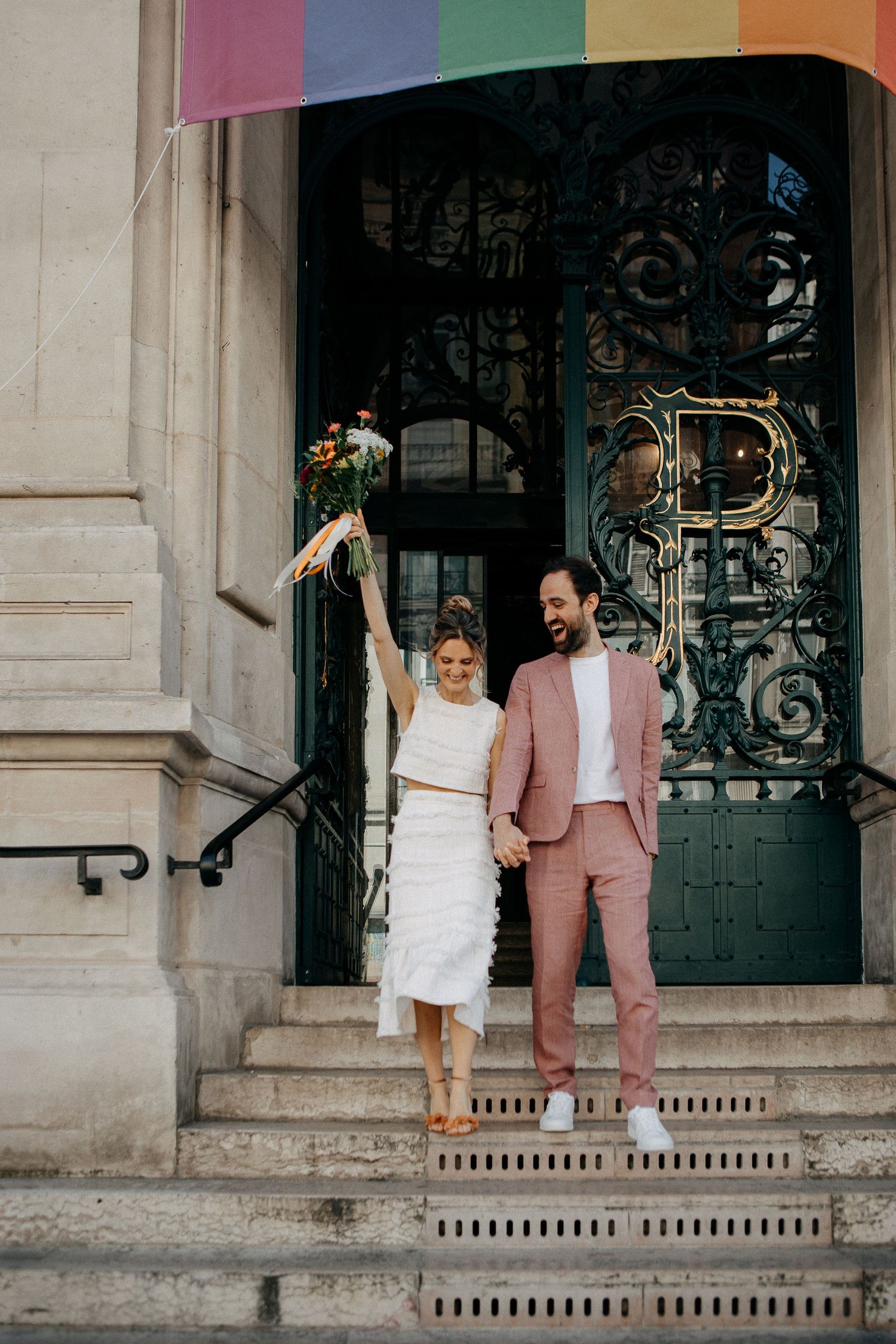 Joyful newlyweds celebrate on steps of ornate building with rainbow flag, bride in white holding bouquet aloft, groom in pink suit.