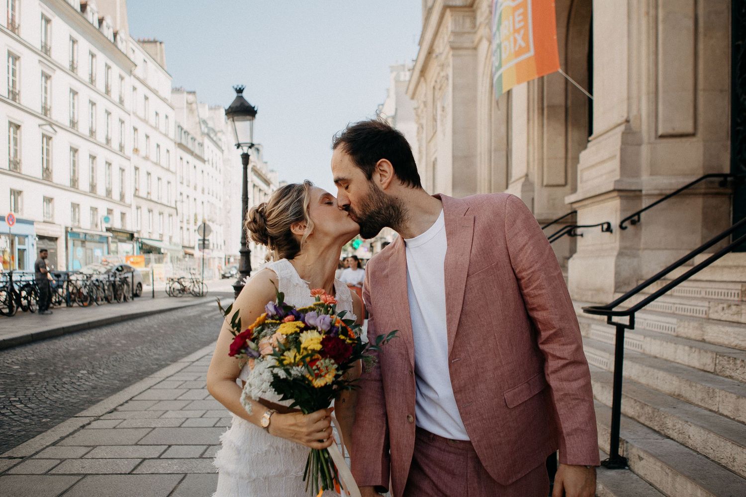 Couple kissing on a Parisian street, bride holding colorful bouquet, groom in pink suit, classic Haussmann buildings backdrop.