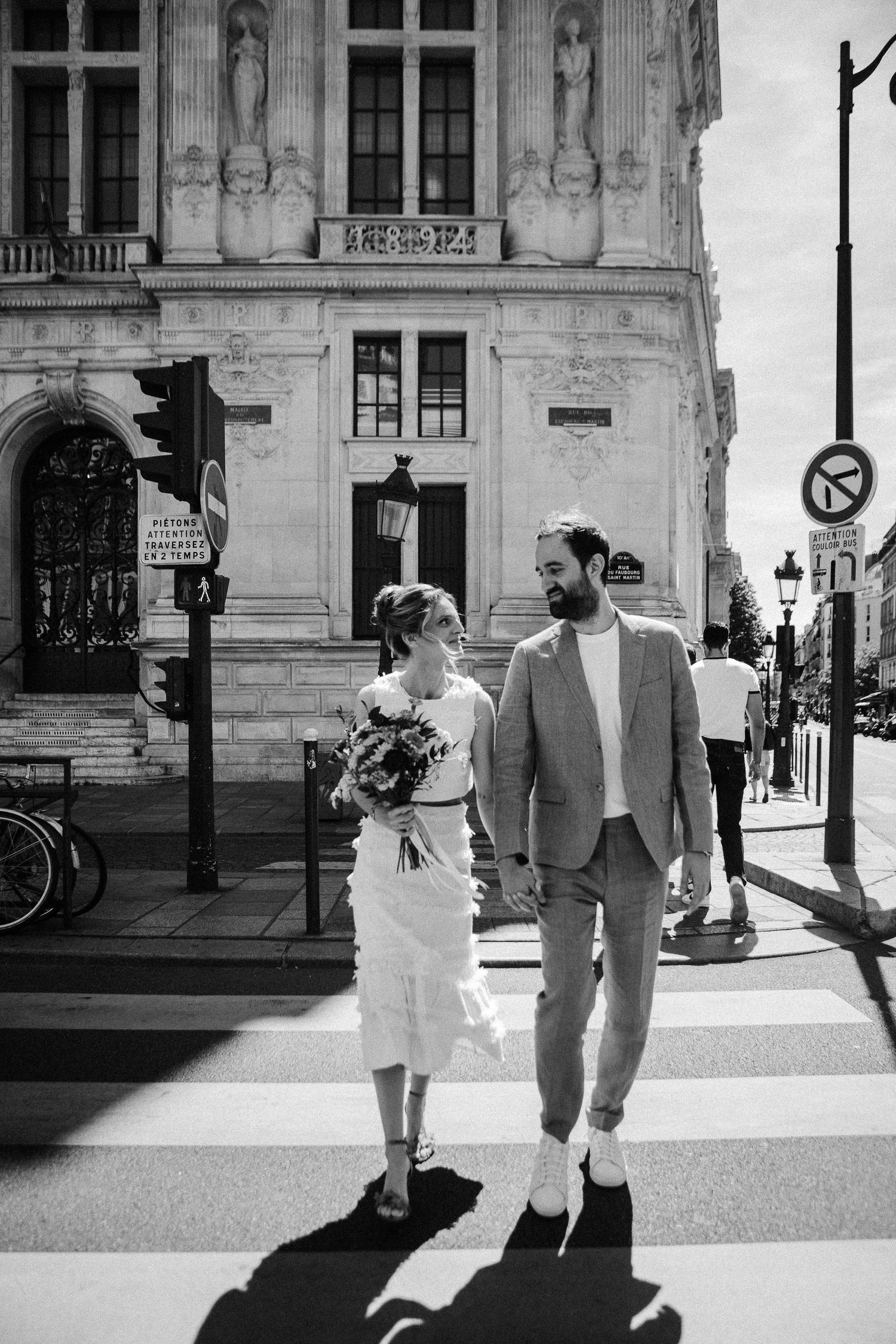 Black and white photo of a couple crossing a street in Paris, woman holding bouquet in white dress, man in suit.