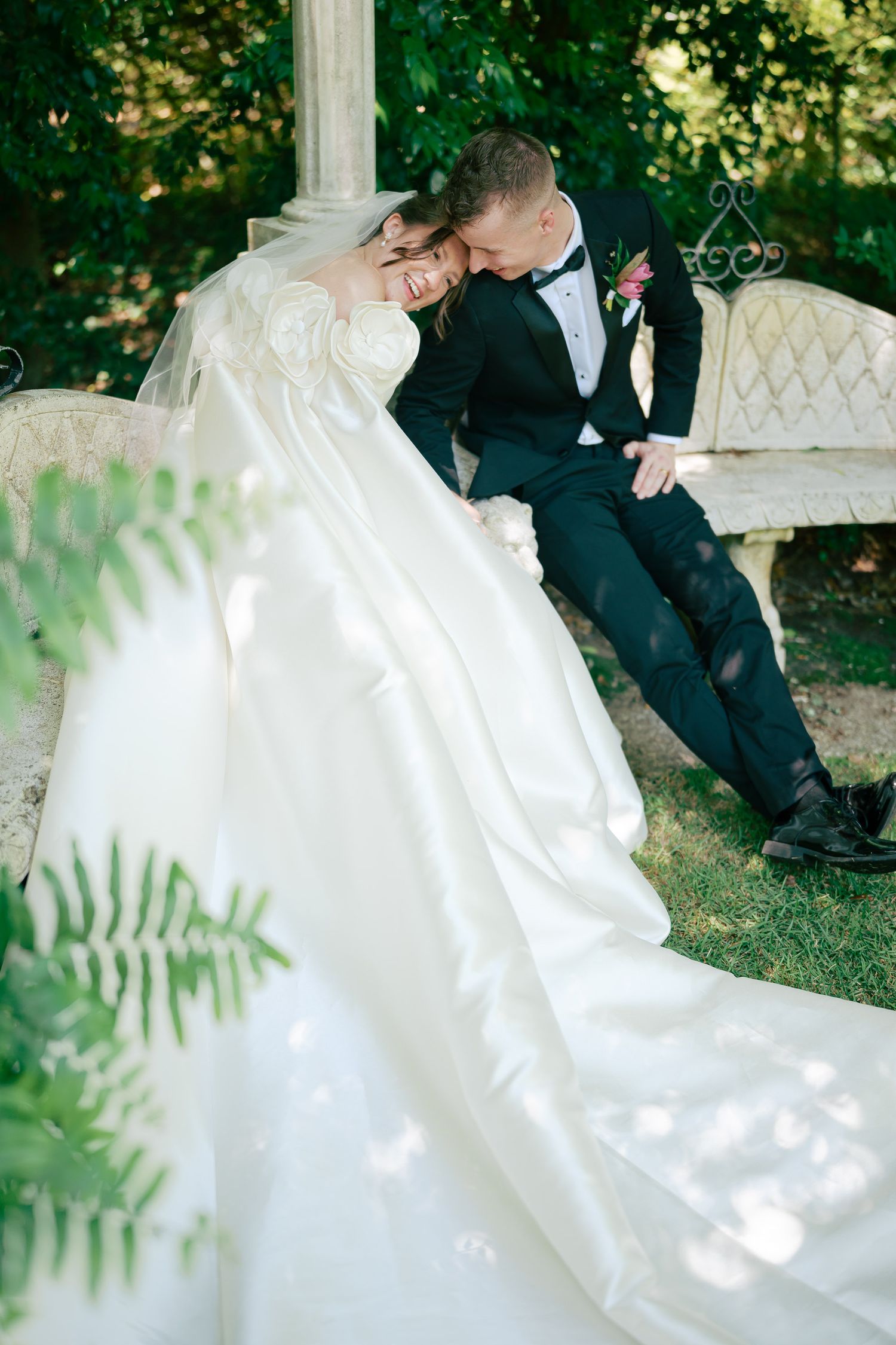 Bride in flowing white gown leans on groom in black tuxedo on garden bench surrounded by lush greenery.