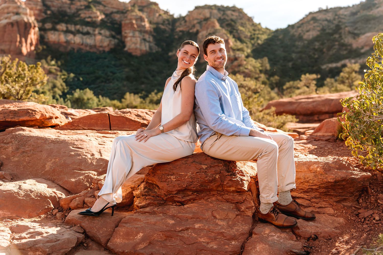 Couple sitting back-to-back on red rocks in Sedona, Arizona, surrounded by lush greenery and stunning red rock formations.