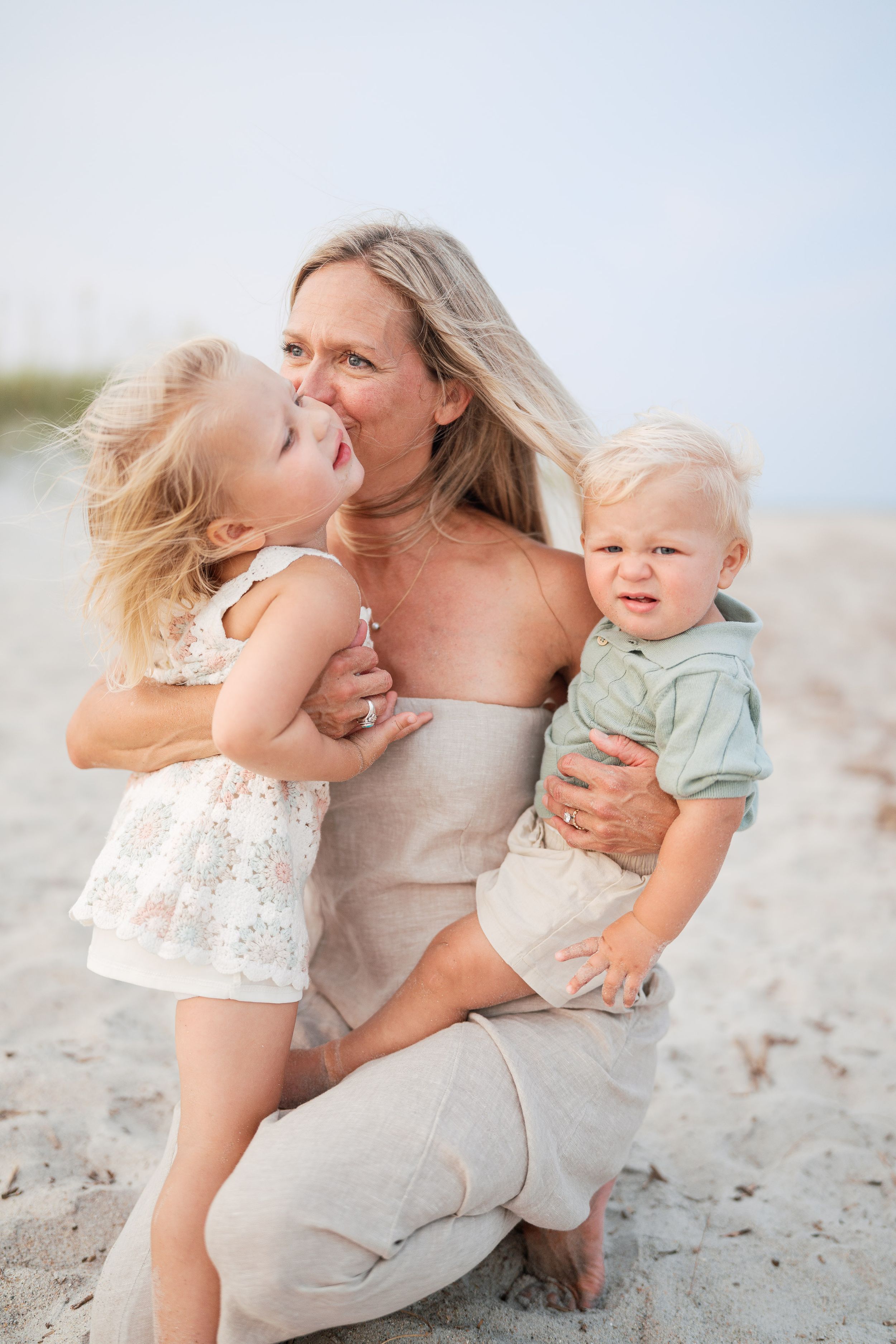 A woman sits on a sandy beach holding two young toddlers, smiling as a little girl gives her a kiss.