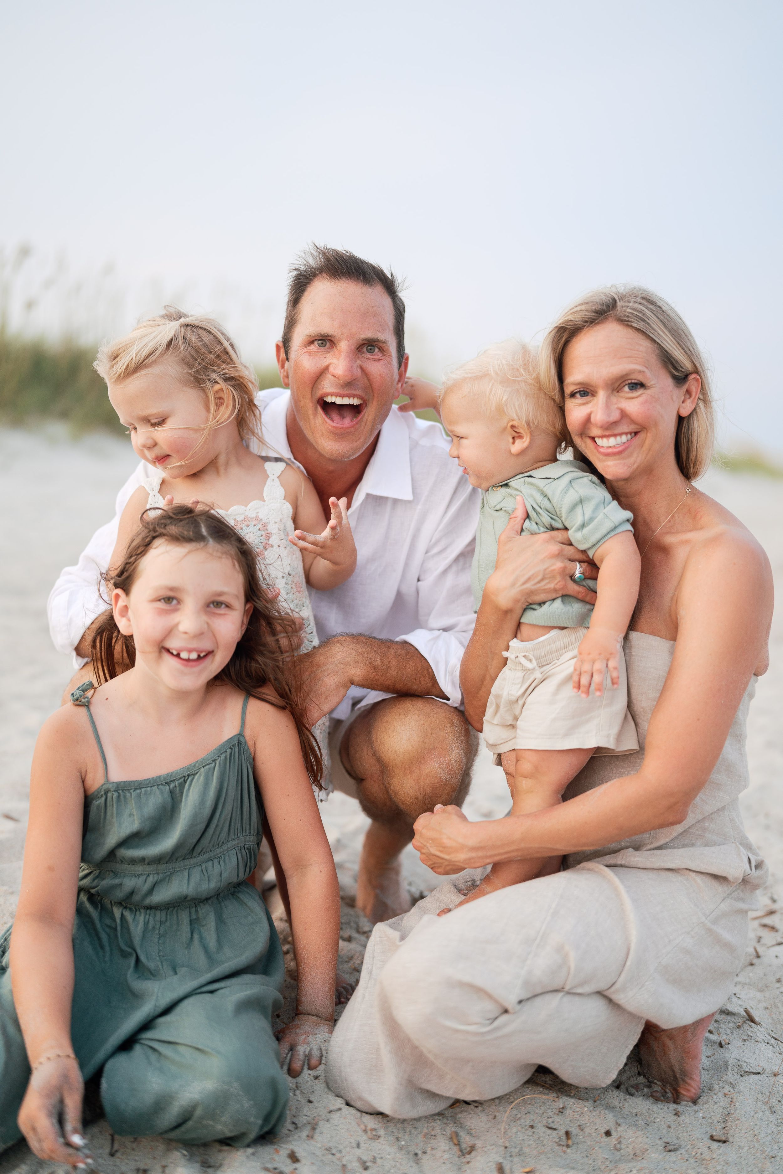 Happy family of five posing together on a sandy beach, parents smiling with three young children in casual summer outfits.