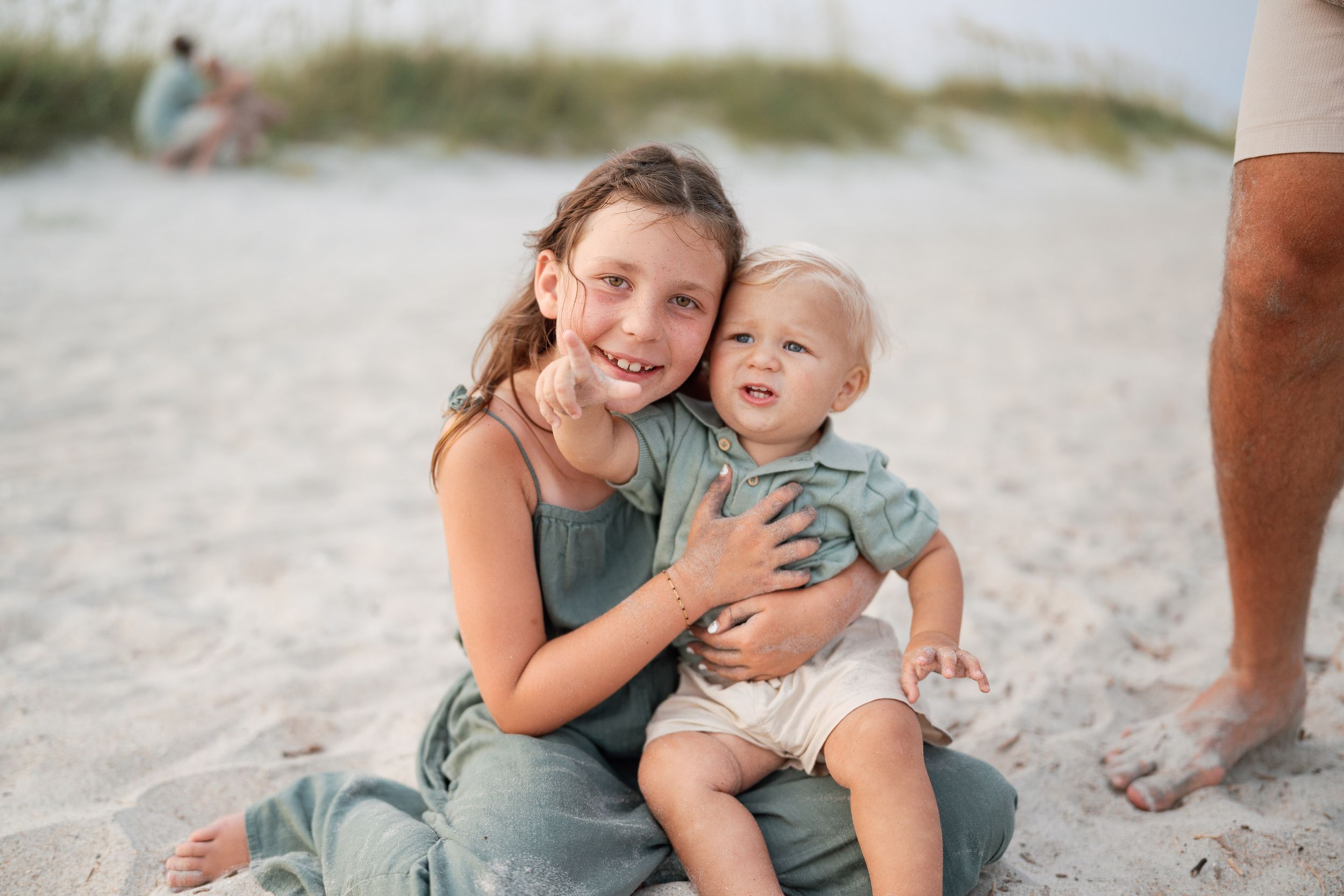 Older sister hugging baby sibling on sandy beach, both wearing sage green outfits, smiling at camera.