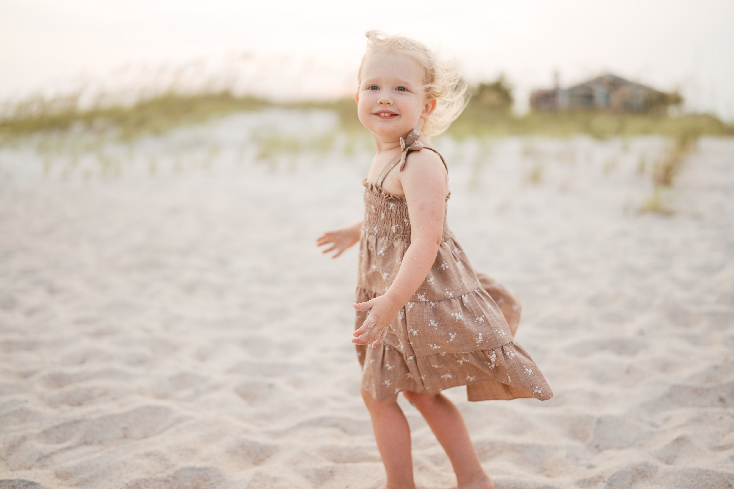 A joyful toddler girl in a brown sundress twirls and plays on a sandy beach at golden hour.