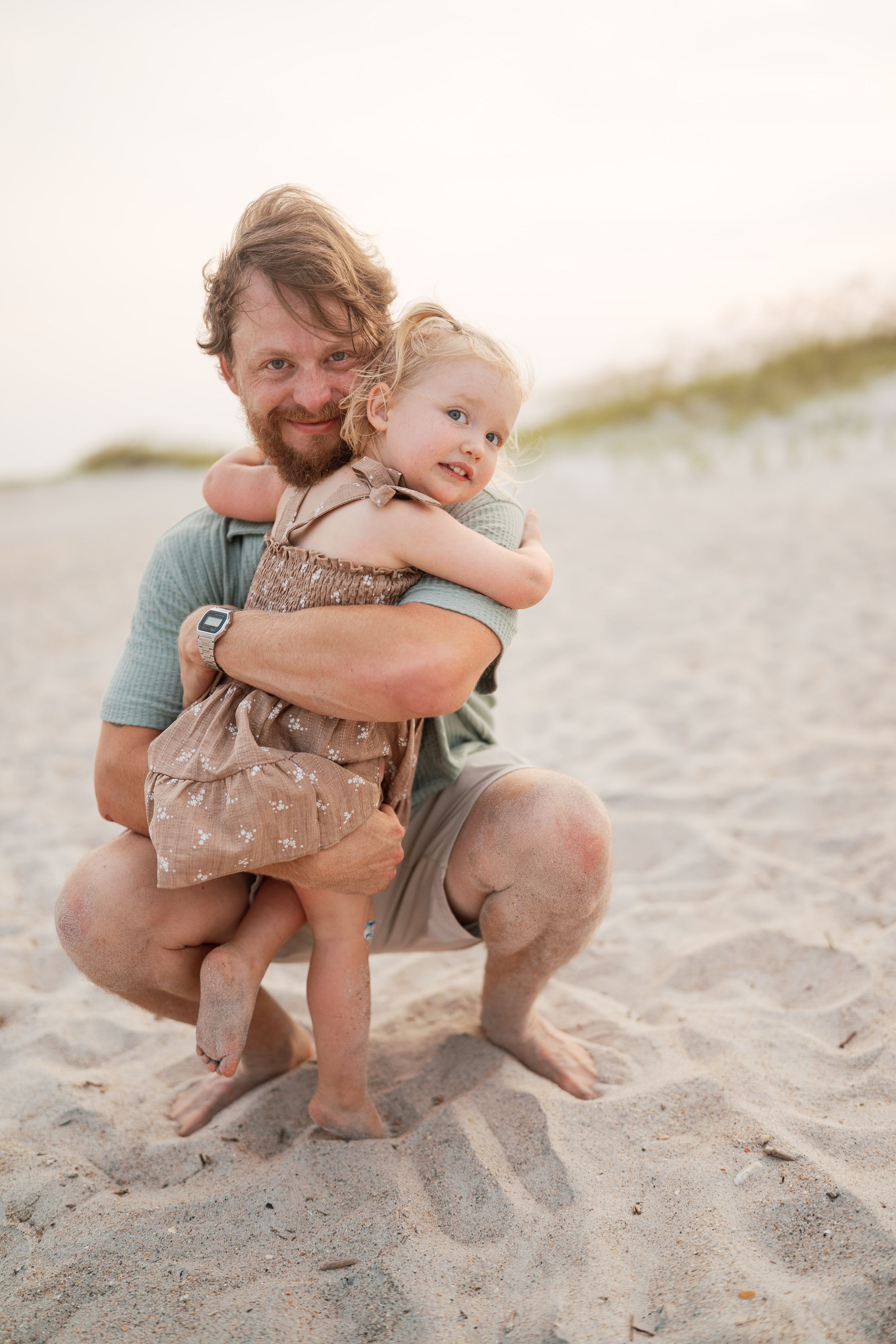 A bearded father crouches on sandy beach hugging his smiling toddler daughter in a floral dress at sunset.