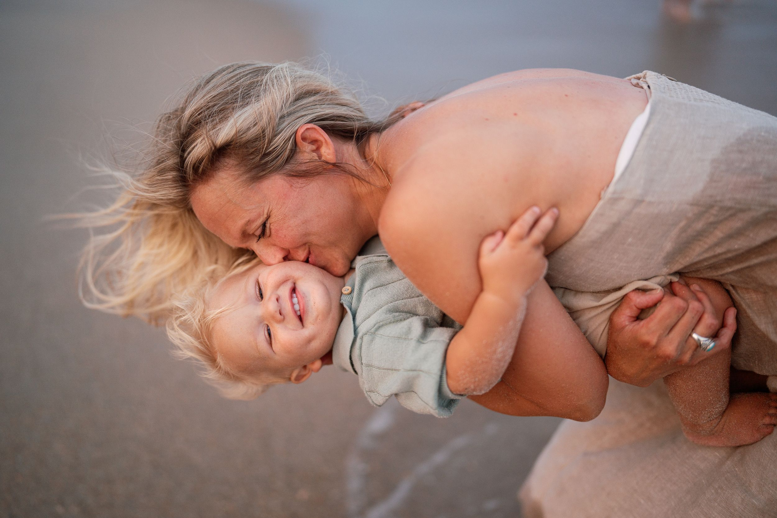A mother joyfully kisses her laughing toddler on a beach, sharing a tender moment by the water.