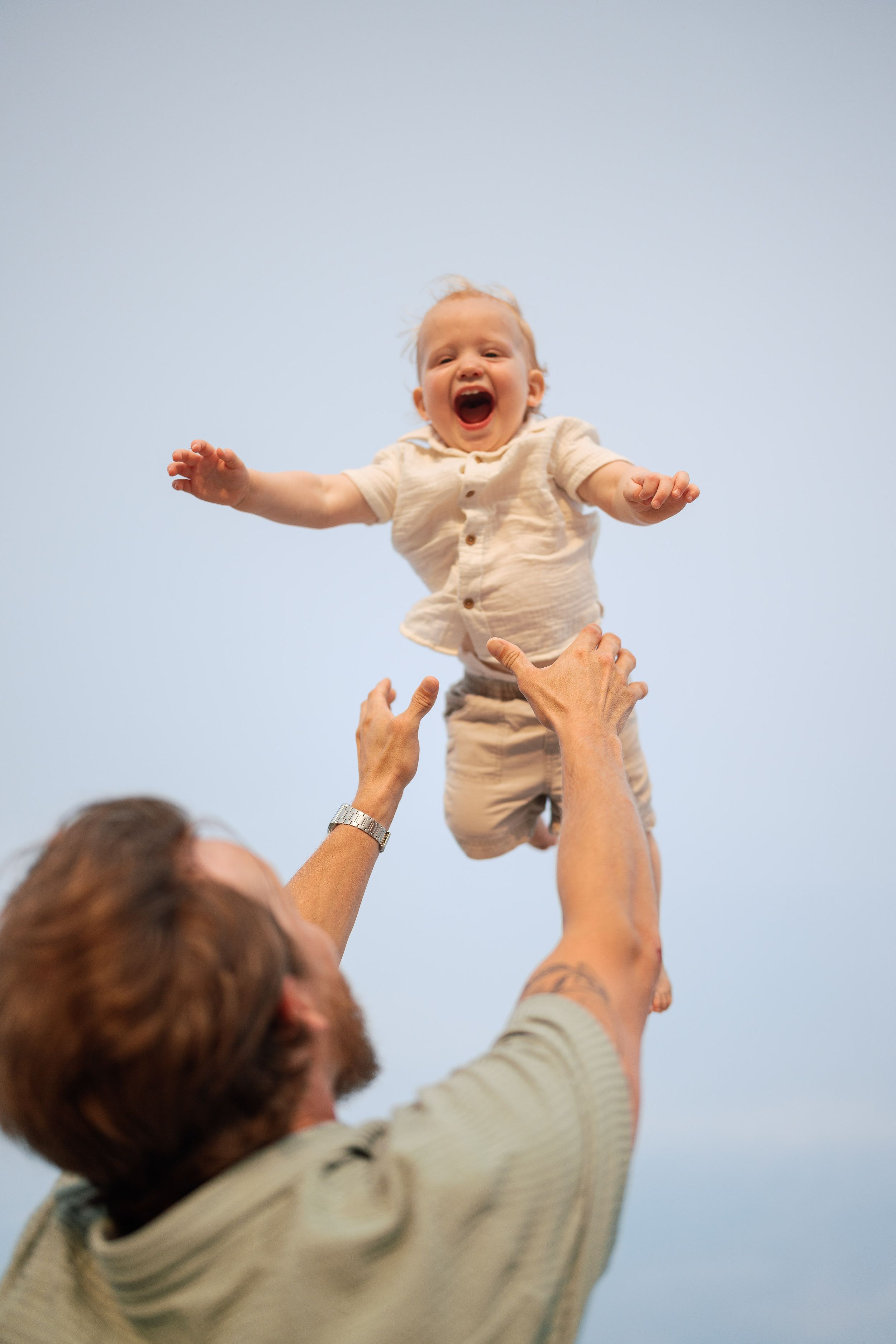 Joyful baby in white outfit laughing as father tosses them in the air against a clear blue sky.