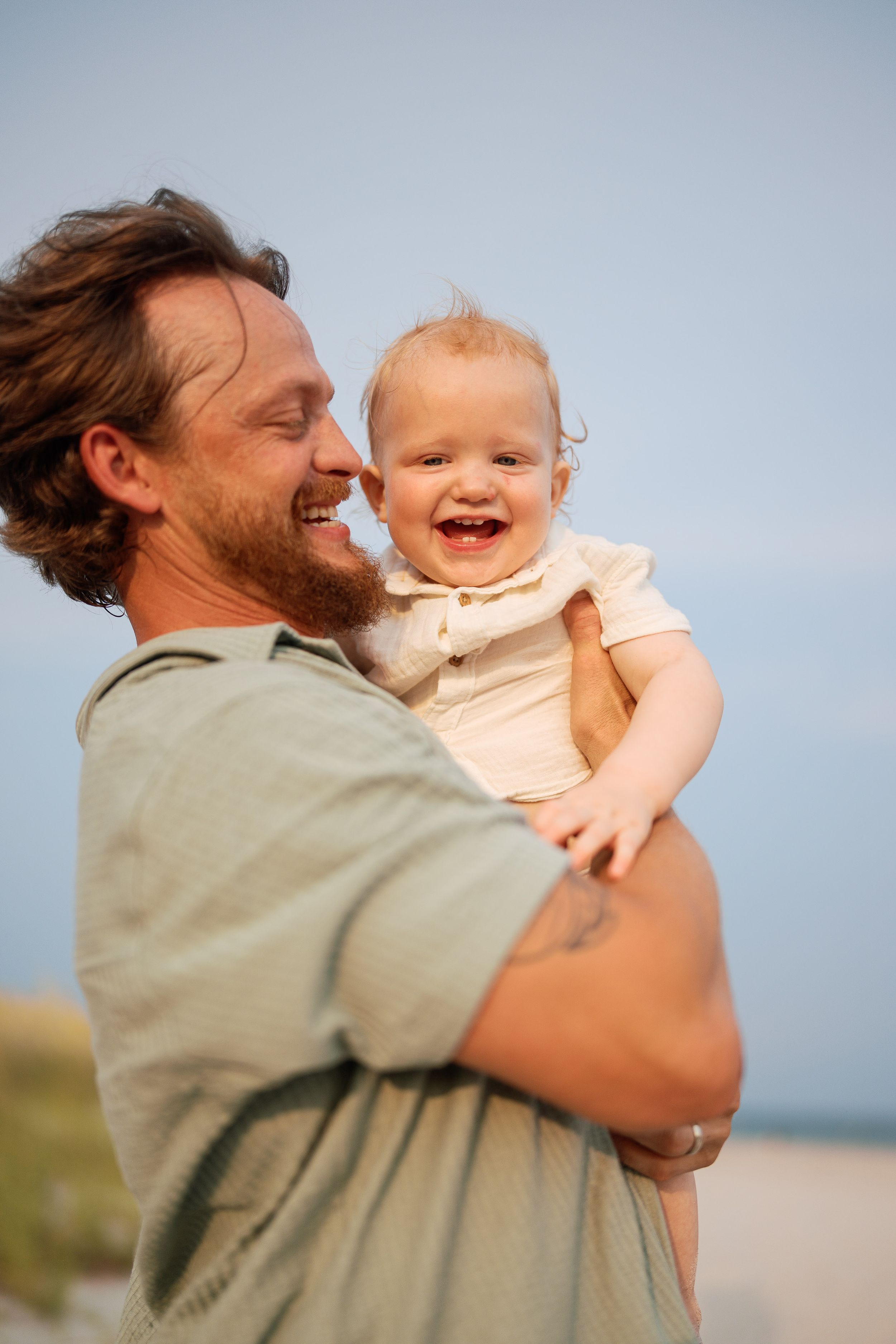 Smiling father holding laughing baby on a sunny beach, sharing a joyful moment outdoors.