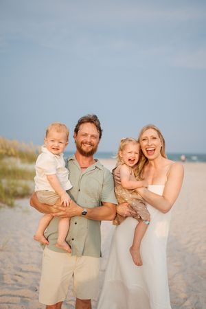 Happy family of four smiling on a sandy beach, parents holding two young children at golden hour.