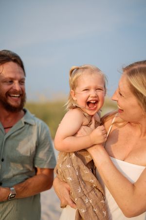Joyful toddler girl laughing while being held by her smiling parents on a sunny beach.