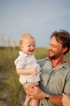 Smiling bearded father holds laughing red-haired baby outdoors near sand dunes on a sunny day.