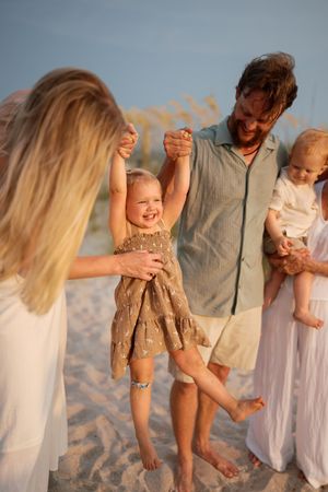 Family playing on sandy beach at sunset, parents lifting laughing toddler while holding baby girl.