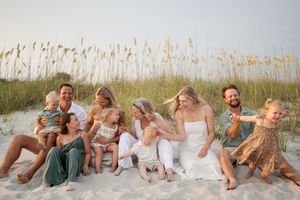 Large family laughing together on sandy beach dunes with sea oats, casual beach portrait session at sunset.