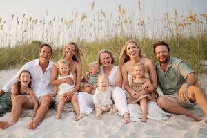 Large family group sitting together on beach sand with sea oats, smiling with babies and children.