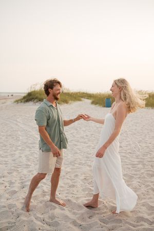 Happy couple dancing barefoot on sandy beach at sunset, woman in white dress holding mans hand.