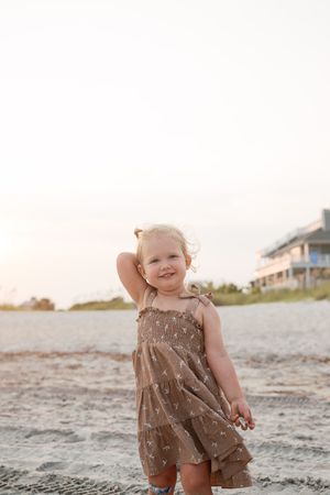 Smiling toddler girl in brown floral dress posing on sandy beach at sunset.