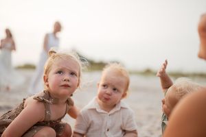 Two young toddlers sit together on a sandy beach, with a girl and boy enjoying outdoor family time.